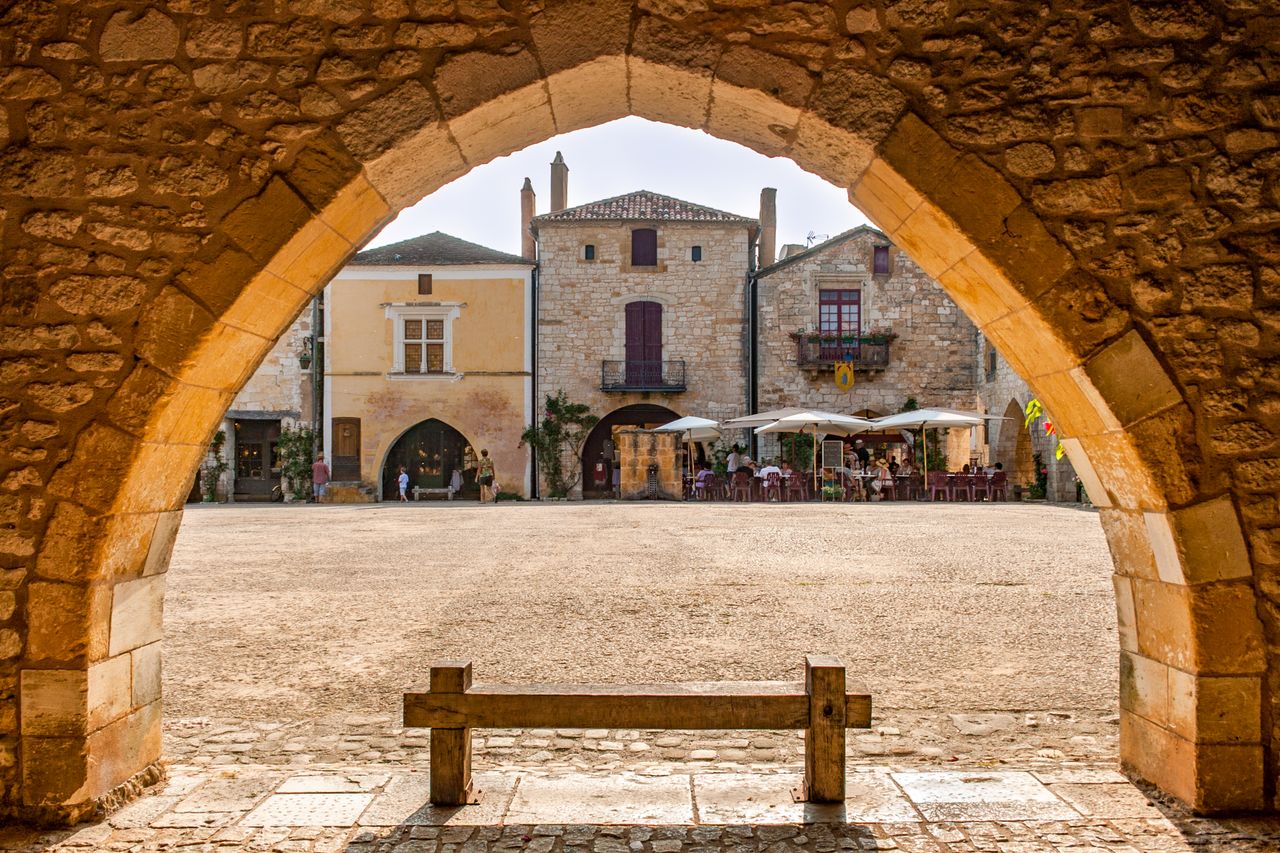 A picturesque view of a medieval square in Monpazier, France captured through a stone archway.
